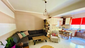 A living room with a sofa, a coffee table, and a dining area with chairs and a table. There is a kitchen in the background with cabinets and a window.