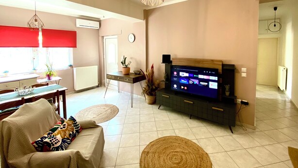 A living room with a TV, a dining area with a table and chairs, and a kitchen area with a window and plants.