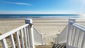 A beach view from a wooden deck with white railings, leading to a sandy beach and ocean. - Cozy, Colorful, Coastal Cottage - Oceanfront 5 bedroom home (Ocean Isle Beach)