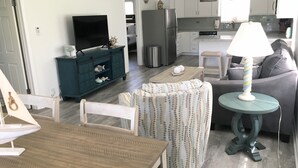 Ein Wohnzimmer mit Holztisch, einer Couch, einem Fernseher und einer Küchenzeile inklusive Kühlschrank und Schränken.