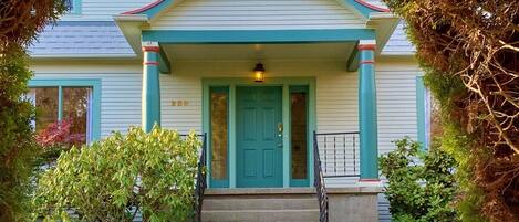 A two-story house with a front porch, a teal door, and a small staircase leading to the entrance.