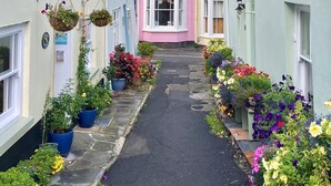 Uma rua estreita com casas de ambos os lados, um edifício rosa no centro e jardineiras com flores penduradas.
