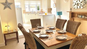 Uma área de refeições com mesa de madeira posta para seis pessoas, rodeada por cadeiras de vime, e uma cozinha ao fundo, além de um objeto decorativo na parede.