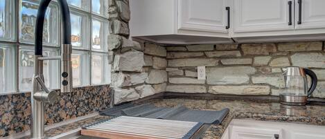 A kitchen with a granite countertop, a stainless steel sink, white cabinets, and a stone wall.
