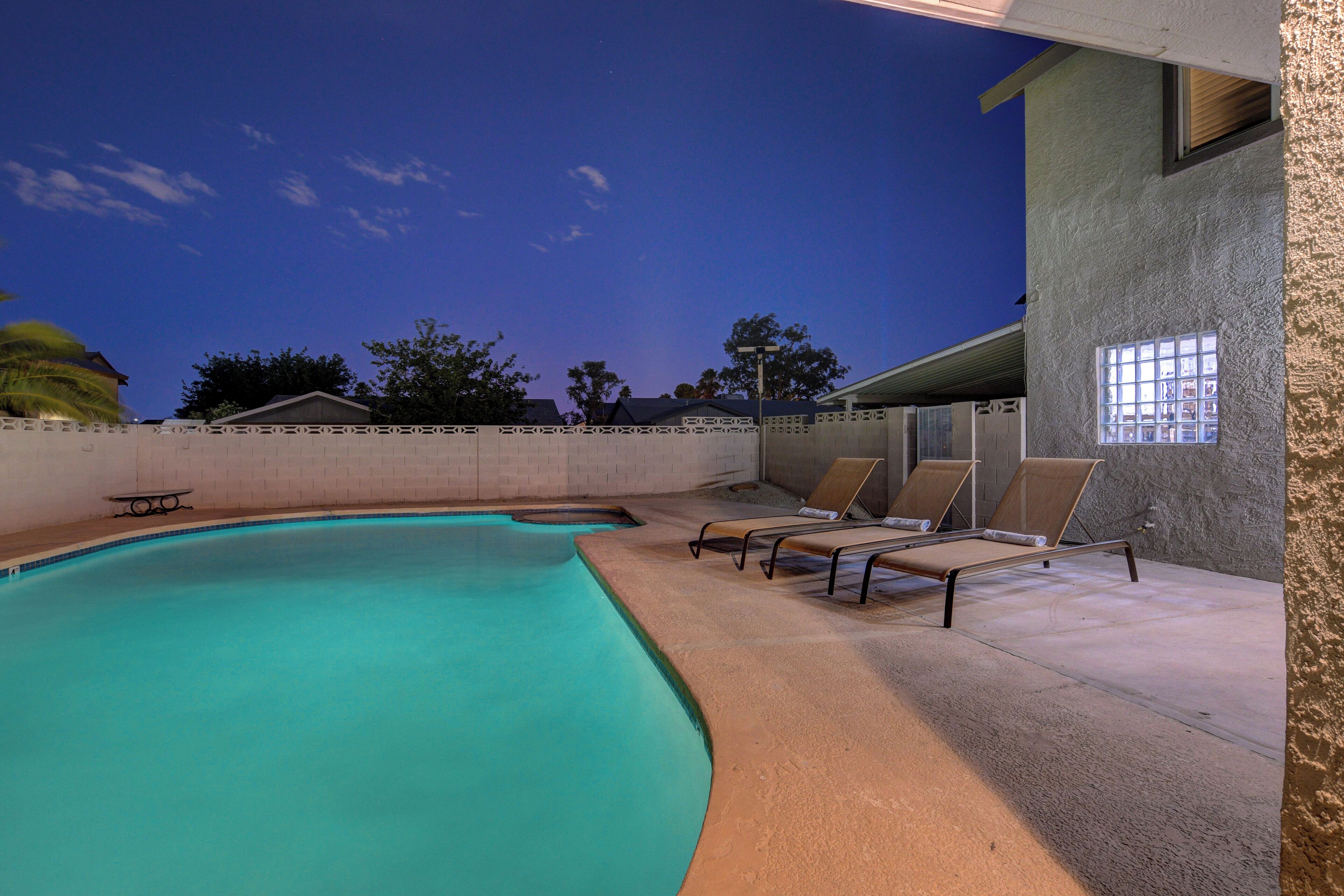 A swimming pool with lounge chairs and a table, surrounded by a concrete wall and a building with a window.