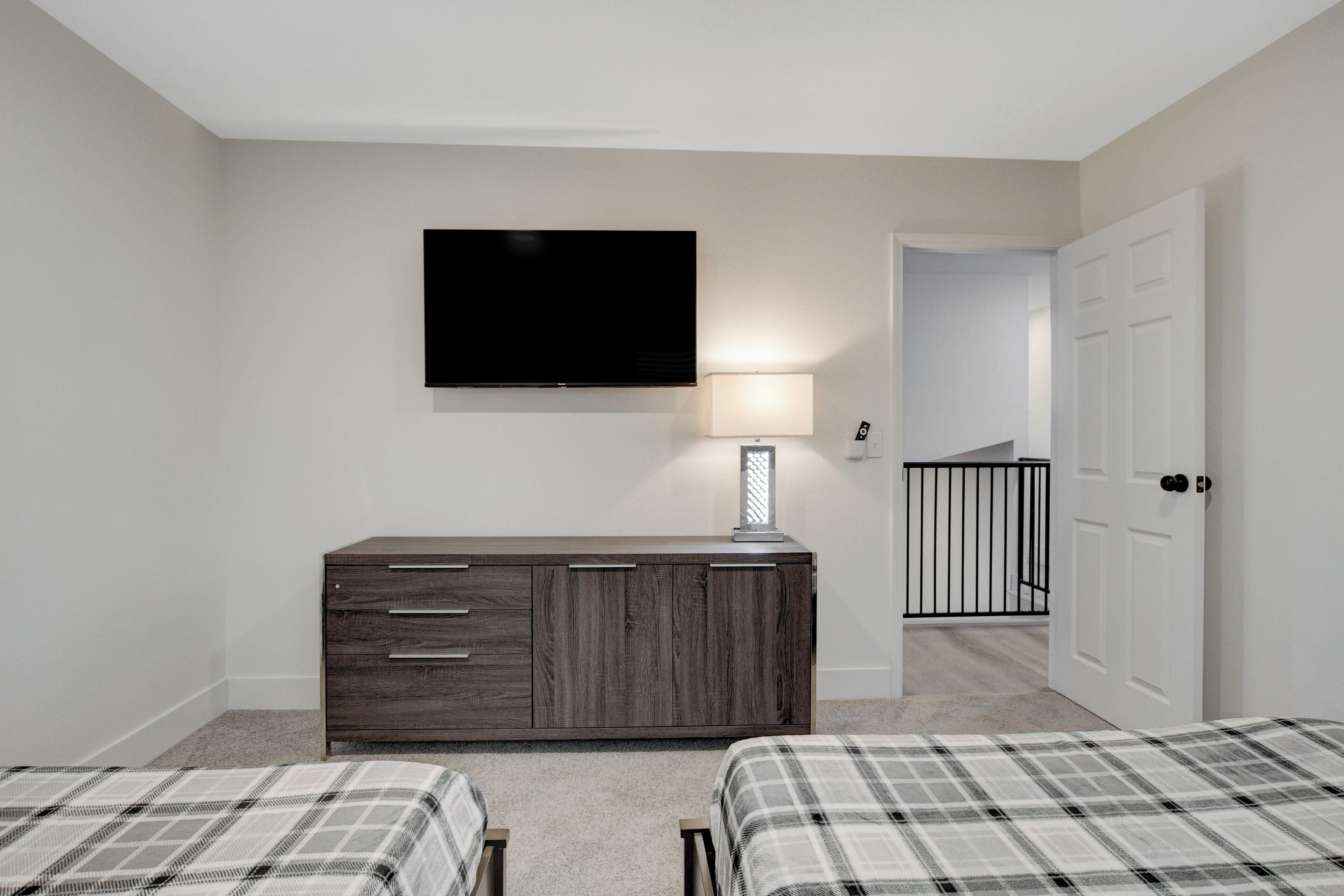 Une chambre avec deux lits, une commode, un téléviseur fixé au mur et une porte donnant sur une autre pièce.