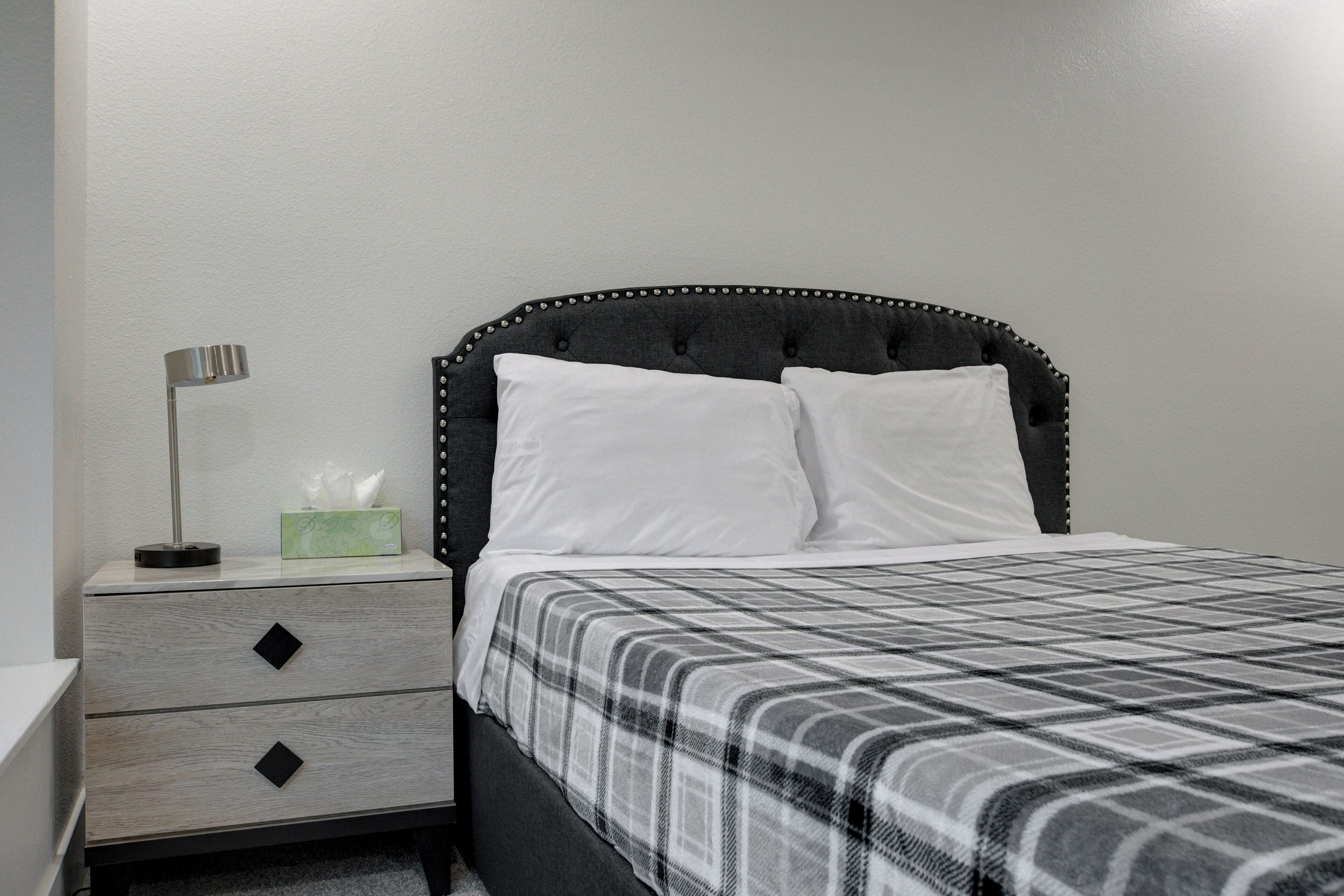 A neatly made bed with a black upholstered headboard, white pillows, and a plaid bedspread. There is a bedside table with a lamp and a tissue box.
