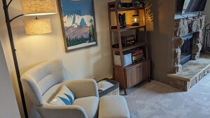 A cozy living room with a large armchair, a wooden bookshelf, a framed picture of mountains, and a stone fireplace. - Peaceful Fraser Condo Retreat, Close to Winter Park! (Fraser)
