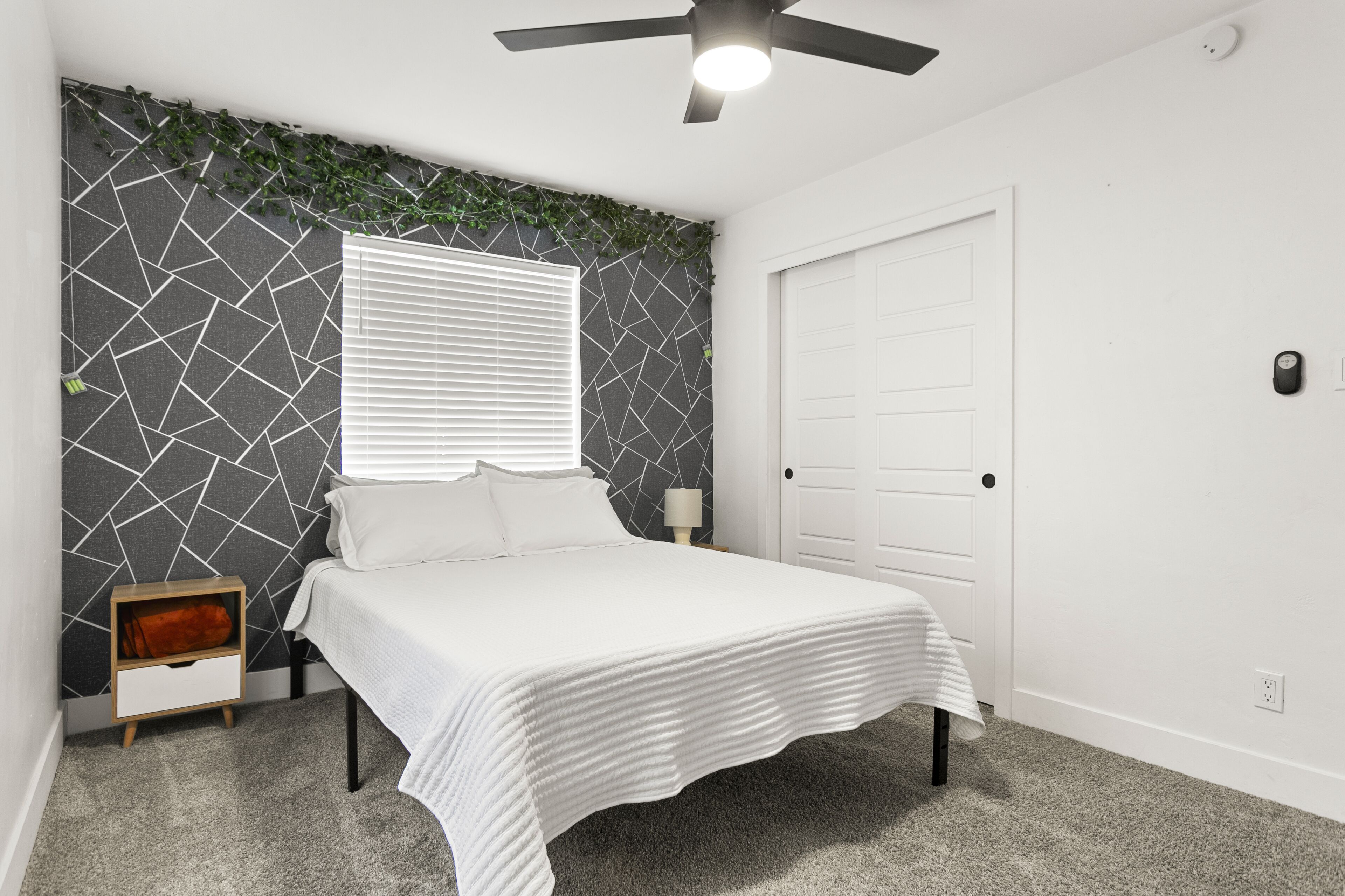 A bedroom with a bed, a nightstand, a wall-mounted lamp, a ceiling fan, and a door.