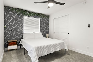 Quarto com cama, mesa de cabeceira, luminária na parede, ventilador de teto e porta.
