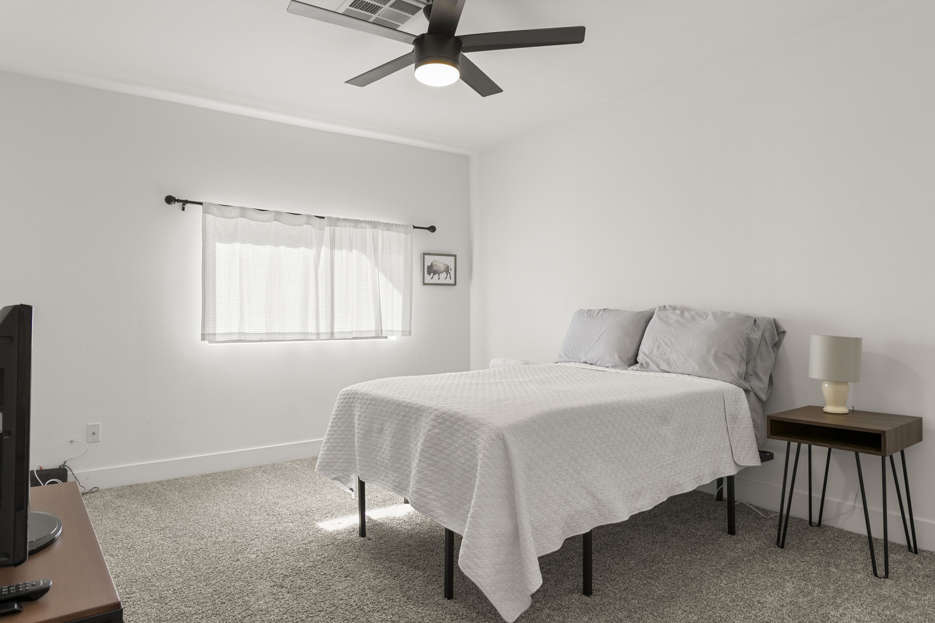 Une chambre à coucher comprenant un lit, une table de chevet, un ventilateur de plafond, une fenêtre avec des rideaux et un tableau accroché au mur.