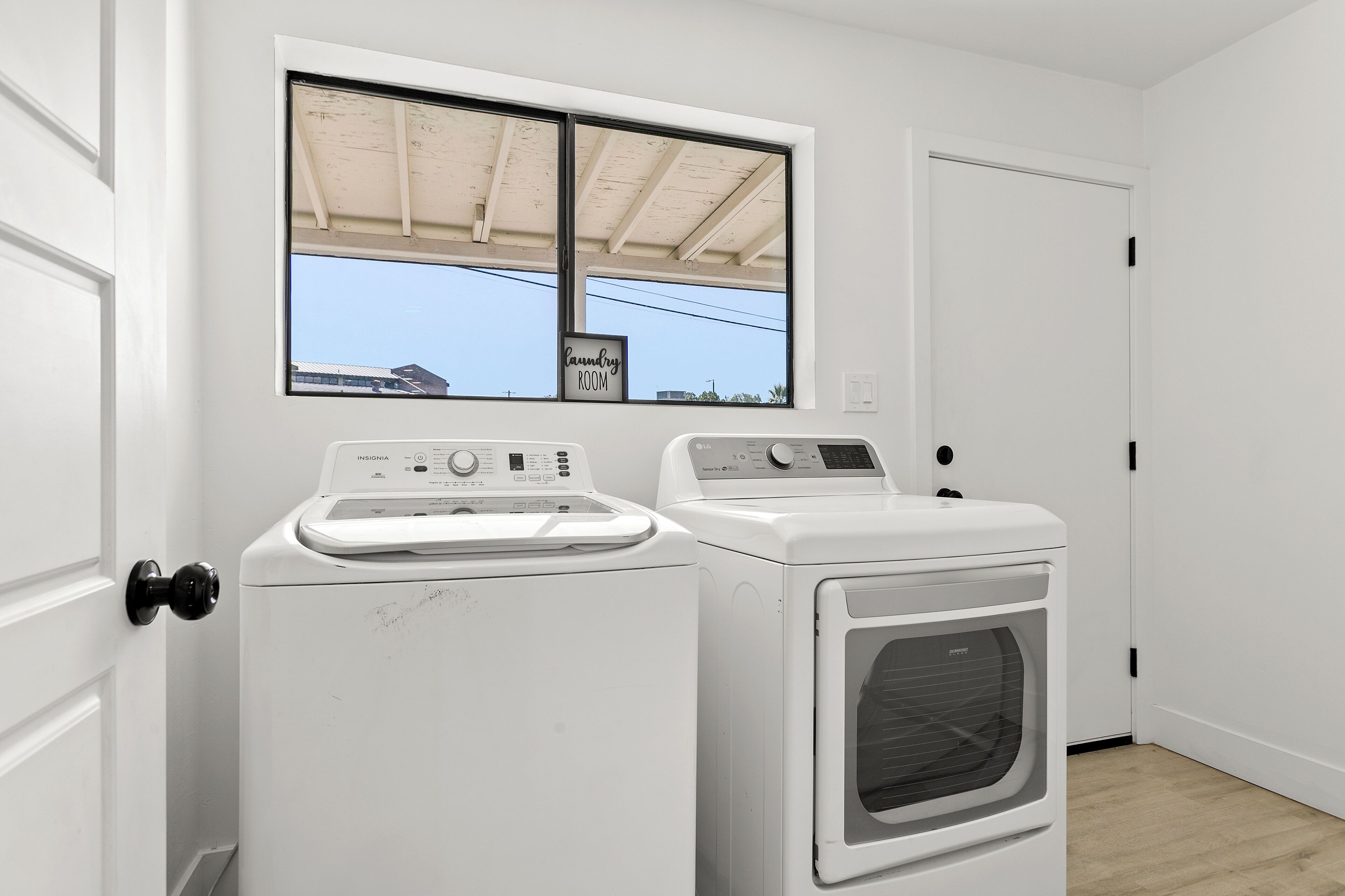 Un cuarto de lavado con dos lavadoras blancas y una secadora, una ventana con vista al cielo, y una puerta.