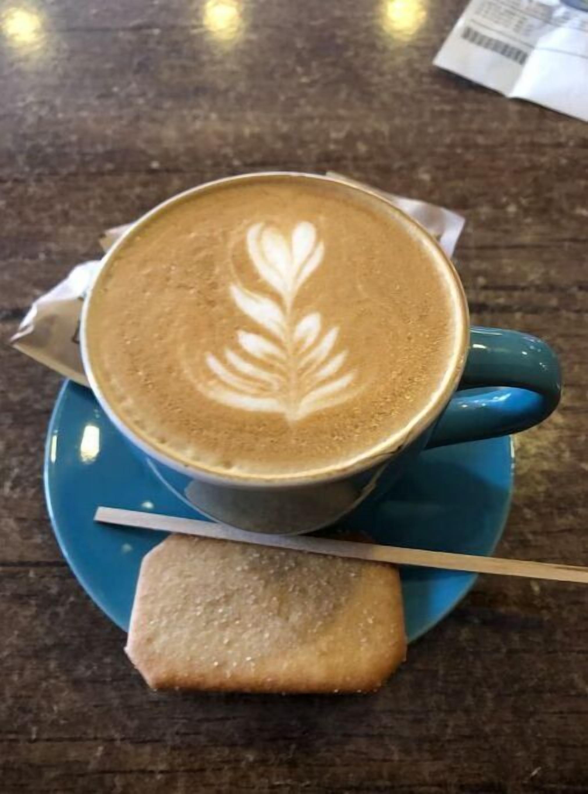 A cup of coffee with latte art on a blue saucer, a spoon, and a biscotti on a wooden table.