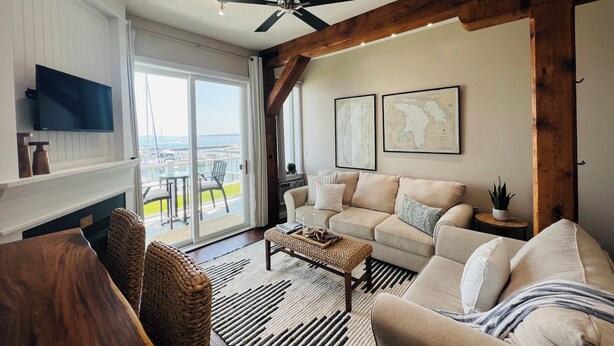 Un vaste salon comprenant un grand canapé, une table basse et un téléviseur fixé au mur. Il y a également un balcon avec vue sur la ville et un ventilateur de plafond.