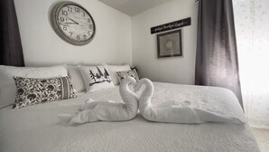 A neatly made bed with a clock and decorative pillows, a framed quote on the wall, and a window with curtains.