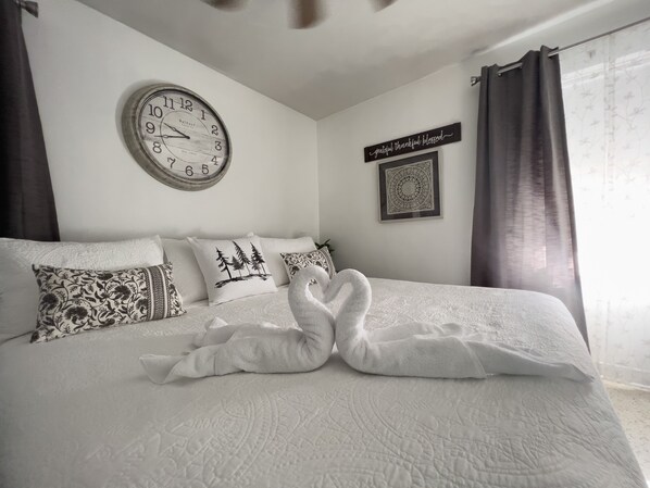 A neatly made bed with a clock and decorative pillows, a framed quote on the wall, and a window with curtains.