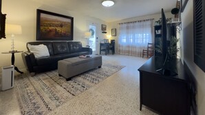 A living room with a black leather sofa, a grey ottoman, a coffee table, a TV stand, and a painting on the wall.
