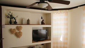 Une chambre à coucher avec un lit, un ventilateur de plafond, un téléviseur posé sur une étagère, des objets de décoration et une fenêtre avec des rideaux.