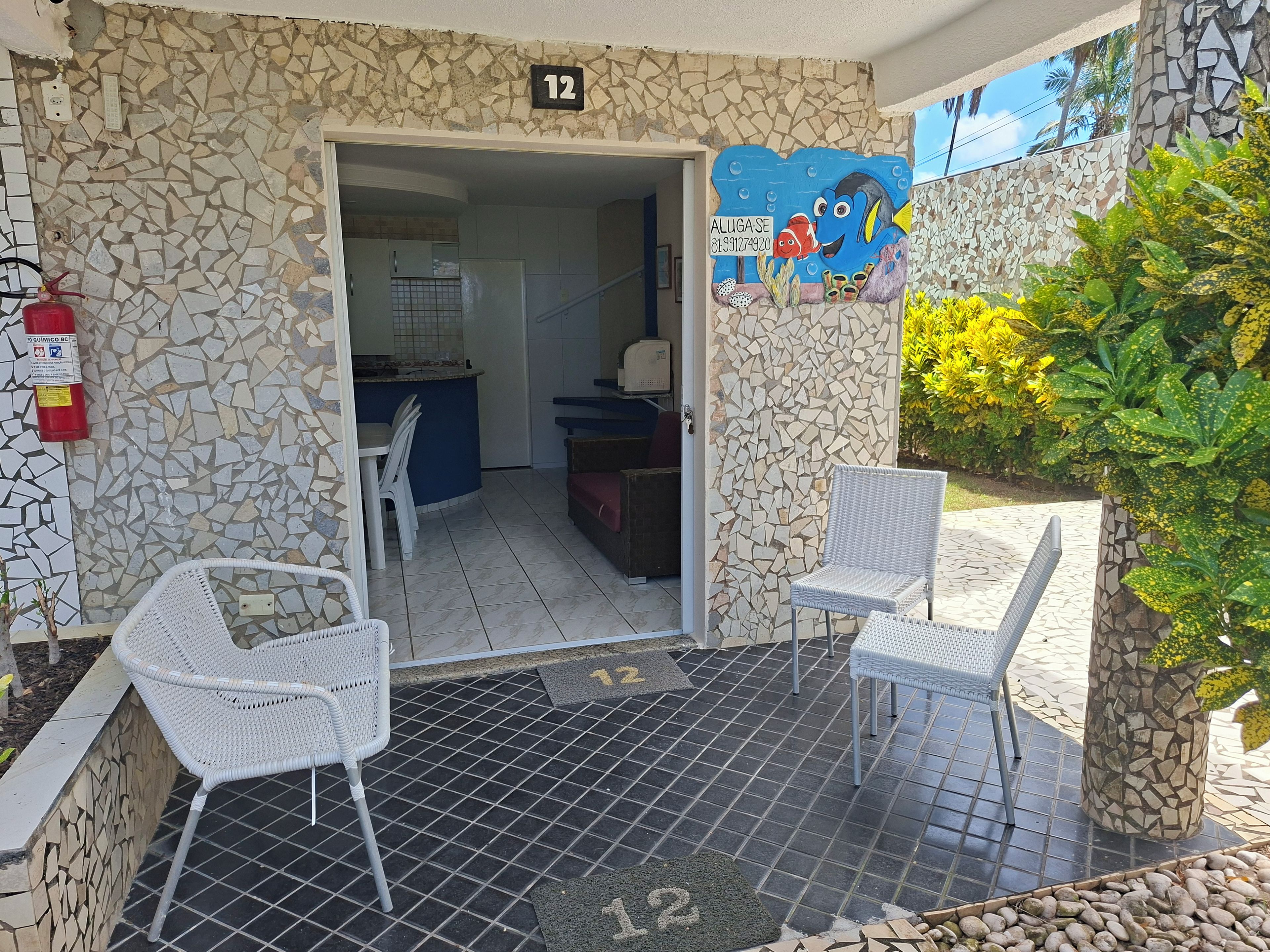 Entrada de hotel con un muro de mosaicos, un cartel con el número 12, un extintor y un pequeño espacio de descanso con dos sillas.