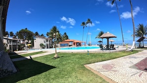 A pool area with a clear blue sky, palm trees, and a building with a red roof. - Chalés Village Gales (Maragogi)