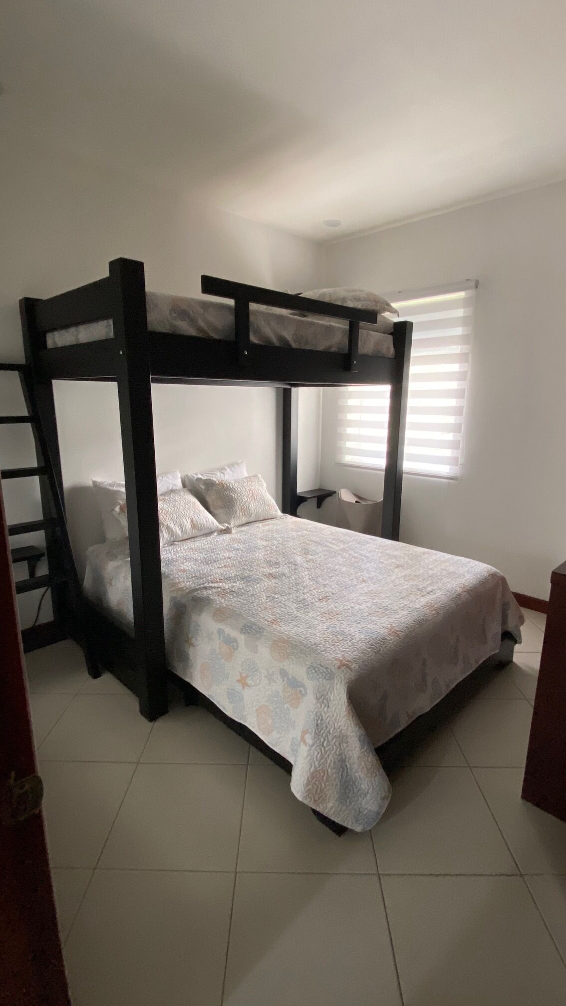 Een slaapkamer met een stapelbed, een wit sprei, een tafeltje en een raam met zonwering.
