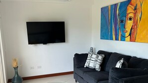 Een woonkamer met een zwarte bank, een gele fauteuil, een glazen salontafel, een plafondventilator, een flatscreen-tv en een kleurrijk abstract schilderij.