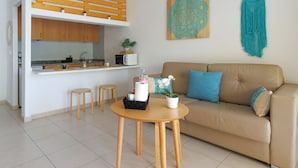 Una sala moderna con un sofá beige, una mesita redonda y una cocina con armarios de madera y encimera blanca.