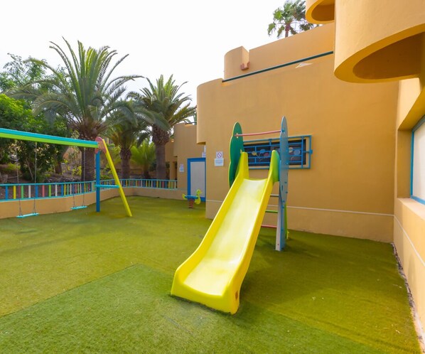 A playground with a yellow slide and swings, surrounded by a building with a yellow exterior and blue accents. - Casa Betta WiFi and Pool iRent Fuerteventura (Corralejo)
