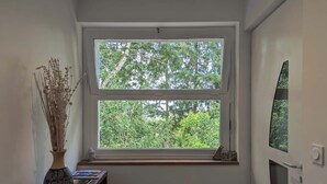 Una habitación con un amplio ventanal que da a un paisaje verde exuberante, un jarrón con plantas secas y un pequeño estante con libros.