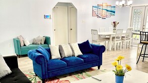 Een woonkamer met een blauwe bank, een zwarte bank, een witte salontafel en een vloerkleed met bloemen. Er is een plafondventilator, een eettafel met stoelen en een deur op de achtergrond.