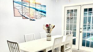 Een eetgedeelte met een witte tafel en stoelen, een kroonluchter en een reeks van drie strandthema schilderijen aan de muur.