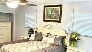 Een slaapkamer met een bed, nachtkastje, plafondventilator, raam met jaloezieën en een ingelijst schilderij aan de muur.