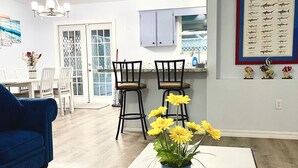 Een woonkamer met een blauwe bank, een witte salontafel met een vaas gele bloemen, en een eethoek met een witte tafel en stoelen.