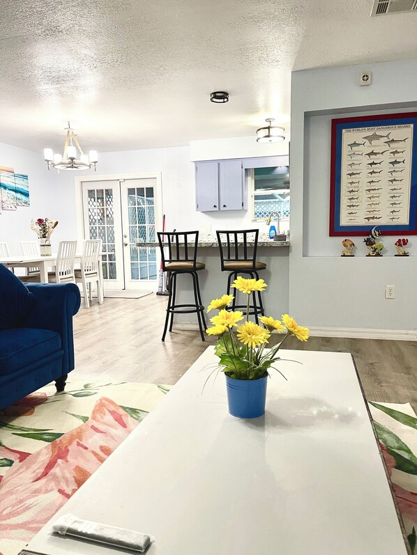 A living room with a blue sofa, a white coffee table with a vase of yellow flowers, and a dining area with a white table and chairs.