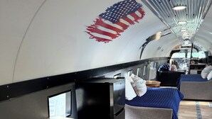 A compact, modern interior with a wooden bench, a small black appliance, and a bed with blue bedding. - Stay in a Piece of History! 1941 WWII Douglas DC-3 Airliner (Deerbrook)