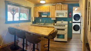 A compact kitchen with wooden cabinets, a microwave, a stove, and a built-in washer and dryer. There are three stools at a bar-style counter. - Abundant Activities for Adventures People (Goldendale)