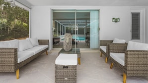 Un espace extérieur moderne, aménagé avec des meubles en osier et une table en verre, offrant une vue sur une piscine et un jardin luxuriant.