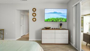 Une chambre à coucher avec un lit, un téléviseur fixé au mur, une commode blanche et une porte donnant sur une autre pièce.