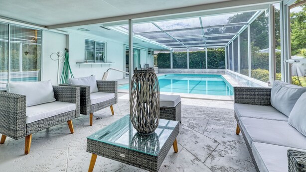 Un espace piscine intérieur moderne, doté de meubles en osier, d’une table basse en verre et offrant une vue sur une piscine extérieure.