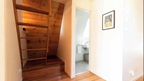 Une pièce avec un plancher en bois, un escalier en bois, une salle de bain avec un lavabo et un miroir, et un tableau encadré accroché au mur.