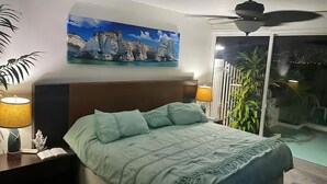 Une chambre à coucher avec un lit, des tables de chevet, un ventilateur de plafond, un tableau et une vue sur l’extérieur.
