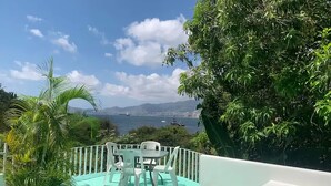 Une piscine turquoise, bordée de chaises blanches et d’une table, entourée d’une végétation luxuriante, d’où l’on peut admirer un plan d’eau et des montagnes lointaines.