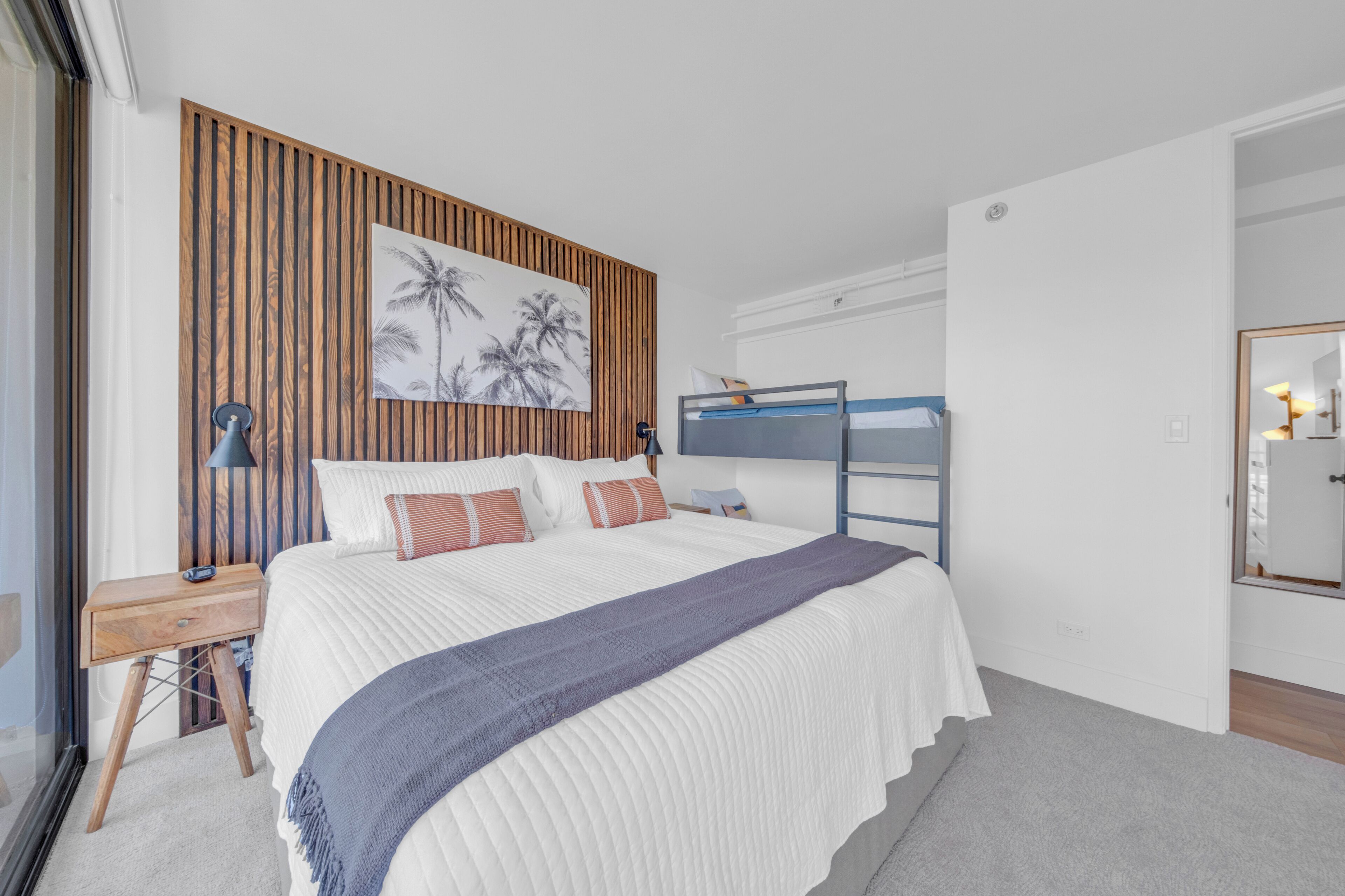 Une chambre avec un lit superposé, un mur recouvert de panneaux de bois, un grand lit avec une literie blanche, une table de chevet avec une lampe et un miroir sur le mur.
