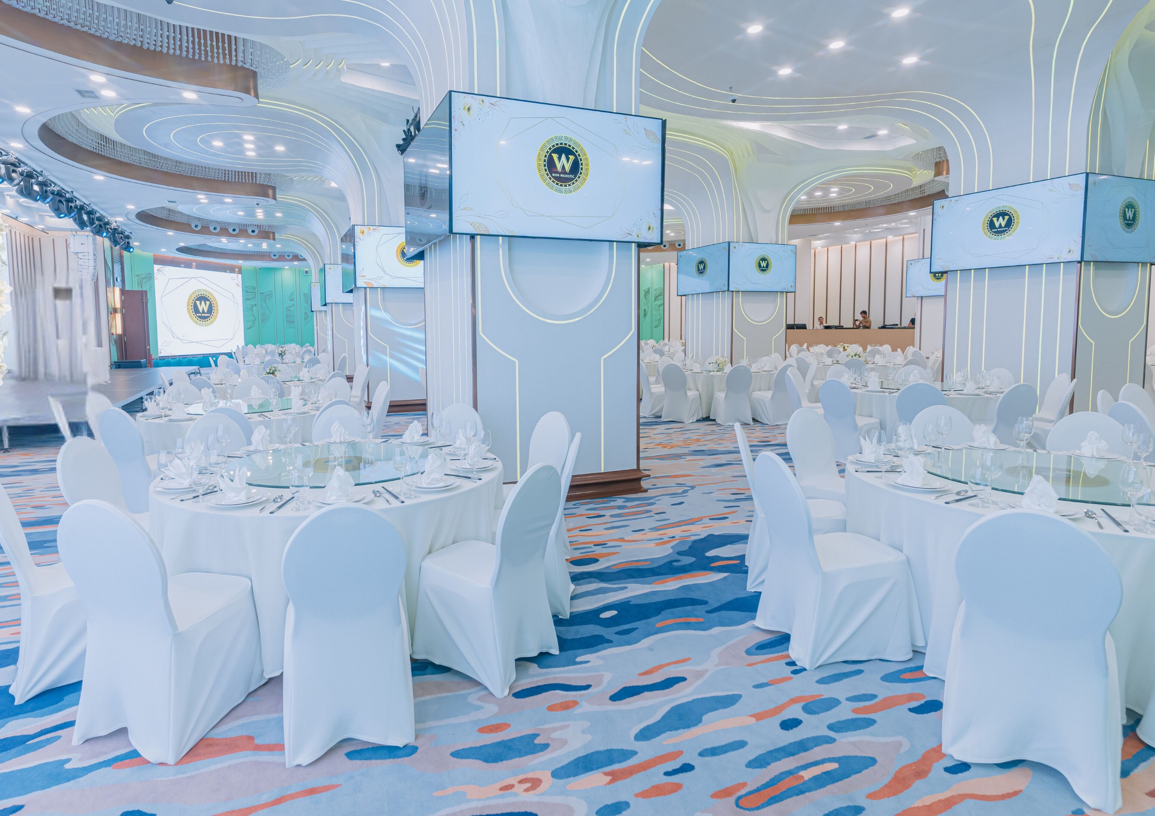 Un salón de banquetes con mesas redondas, sillas blancas y una alfombra azul.