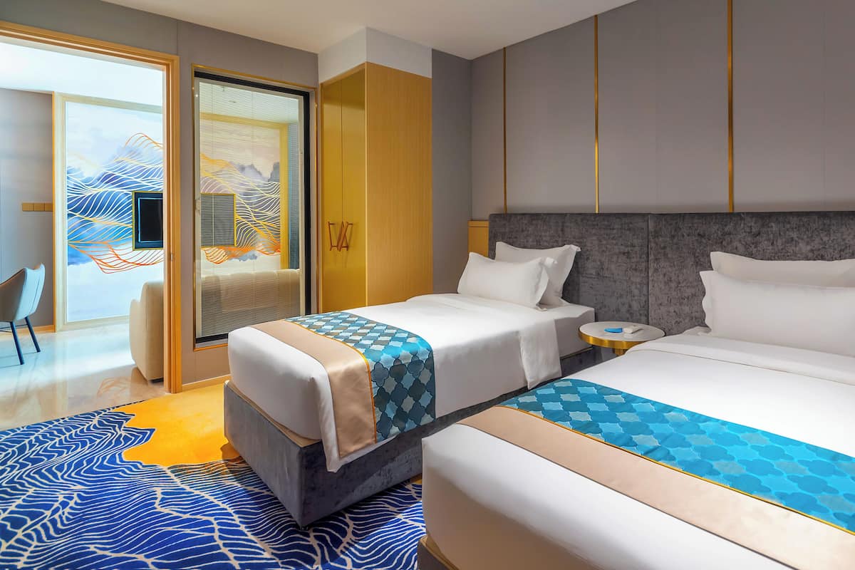 A hotel room with two beds, a large window with a view, a seating area with a chair, and a decorative wall panel.