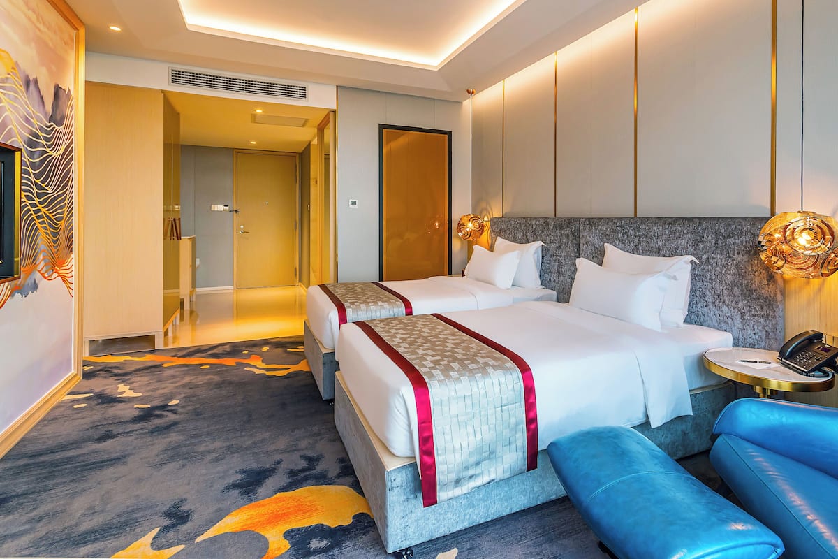 A hotel room with two beds, a blue armchair, a desk with a phone, and a decorative wall.