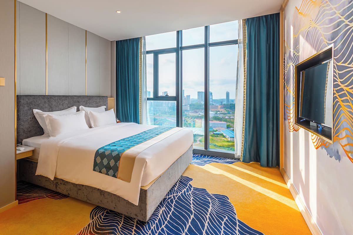 A modern hotel room with a large bed, a city view, and a flat-screen TV mounted on the wall.