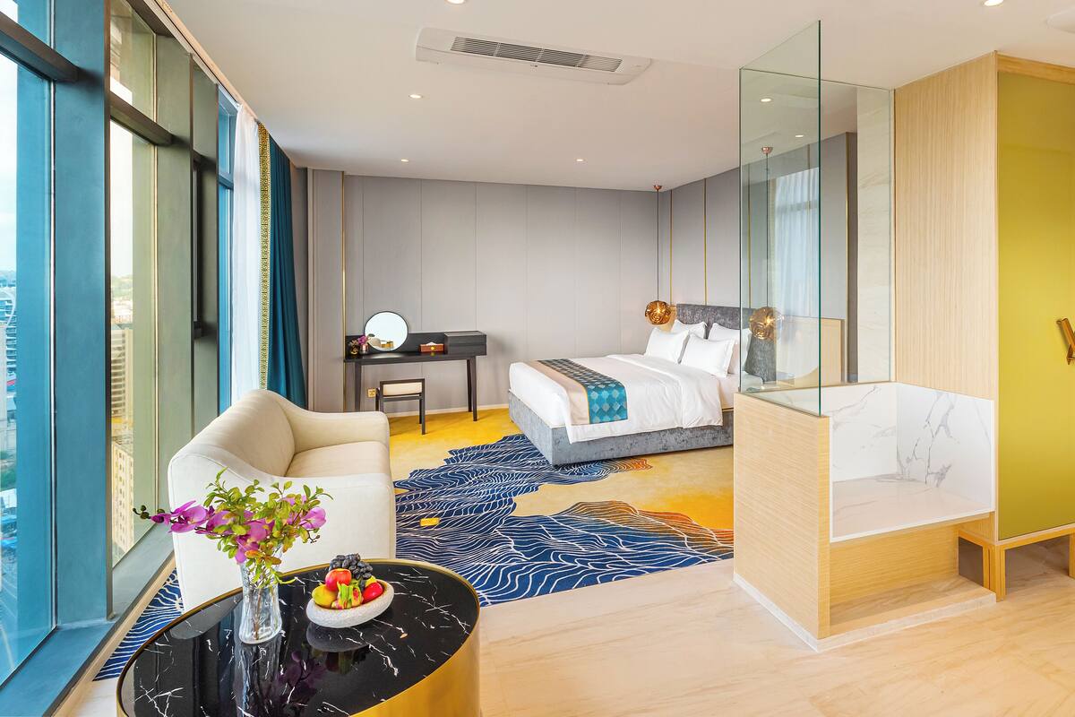 A modern hotel room with a bed, a desk, a chair, a small table with a vase of flowers, and a view of the city through large windows.