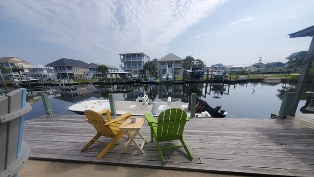 A wooden dock with two Adirondack chairs and a table, overlooking a marina with boats and houses in the background. - Deep-Water Canal Front Coastal Cottage Only Steps to the Beach & Pet Friendly!  (Surf City)