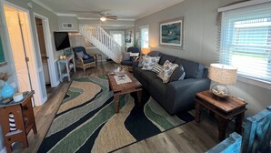 A spacious living room with a dark grey sofa, a wooden coffee table, and a patterned rug. A staircase leads to another level, and a TV is mounted on the wall. - Deep-Water Canal Front Coastal Cottage Only Steps to the Beach & Pet Friendly!  (Surf City)